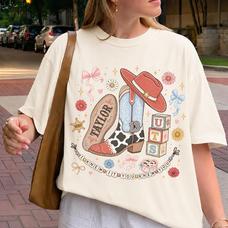 Pode incluir: Uma t-shirt cor de creme com uma impress&atilde;o gr&aacute;fica de uma bota de cowboy com padr&atilde;o de vaca, um chap&eacute;u de cowboy vermelho, blocos de alfabeto com as letras UTS, uma estrela de xerife, la&ccedil;os, flores e o texto TAYLOR e I KNEW IT YOU KNEW YOU.