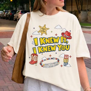 Puede incluir: Una camiseta color crema con un estampado gr&aacute;fico que incluye el texto "I KNEW IT, I KNEW YOU". El dise&ntilde;o presenta una estrella de sheriff amarilla, un sombrero de vaquero rojo, botas vaqueras rojas, una pulsera de cuentas y bloques.