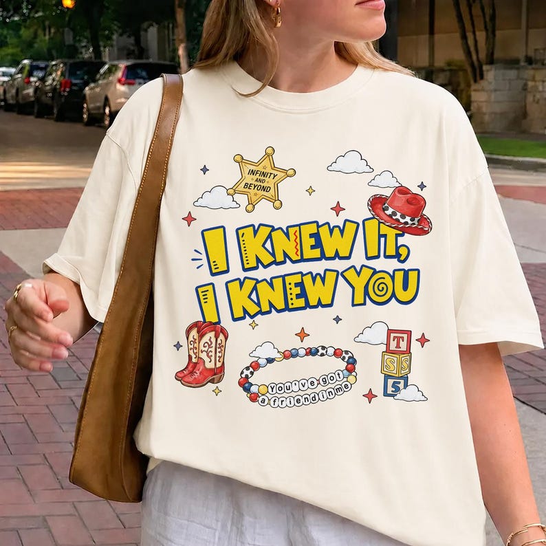 Puede incluir: Una camiseta color crema con un estampado gr&aacute;fico que incluye el texto "I KNEW IT, I KNEW YOU". El dise&ntilde;o presenta una estrella de sheriff amarilla, un sombrero de vaquero rojo, botas vaqueras rojas, una pulsera de cuentas y bloques.