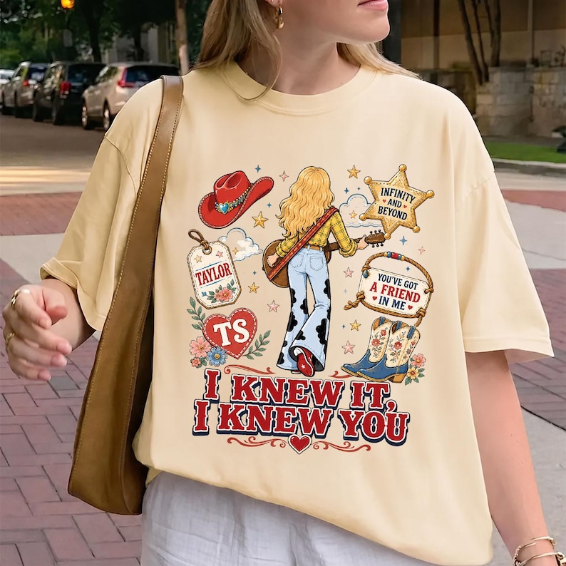 May include: A cream-colored t-shirt featuring a graphic print of a person with long blonde hair, a red cowboy hat, a guitar, and cow-print pants. The design includes a sheriff star, boots, and text reading "TAYLOR", "TS", "INFINITY AND BEYOND", "YOU'VE GOT A FRIEND IN ME", "I KNEW IT, I KNEW YOU".