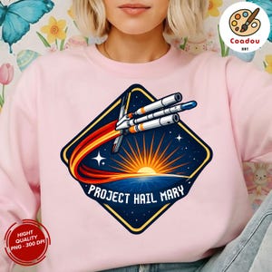 Può includere: Felpa rosa chiaro con una grafica a forma di diamante blu navy raffigurante un razzo, la scritta "PROJECT HAIL MARY" e un sole che sorge su un orizzonte blu. La felpa ha un girocollo.