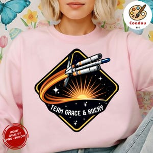 Può includere: Felpa rosa chiaro con una grafica di un razzo spaziale nero, giallo e arancione. La grafica include il testo "TEAM GRACE & ROCKY" e un motivo a raggiera. La felpa ha un girocollo.