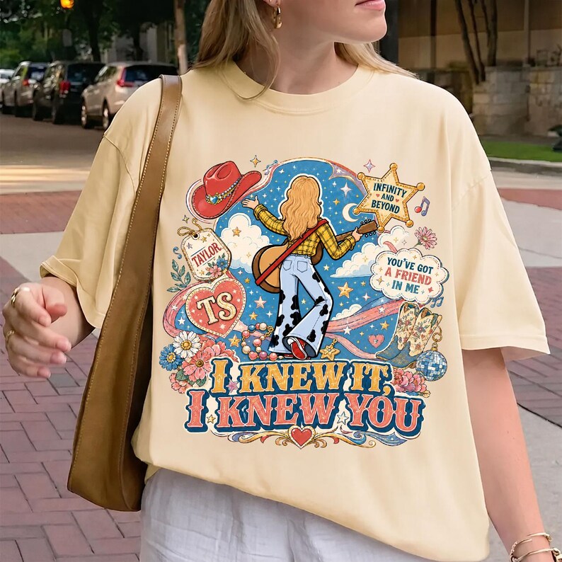 May include: A cream-colored t-shirt featuring a colorful graphic print of a person with long blonde hair playing a guitar, surrounded by a red cowboy hat, a star, flowers, and text that reads "Taylor", "TS", "Infinity and Beyond", "You've Got a Friend in Me", and "I Knew It, I Knew You".