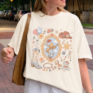 May include: A cream-colored short-sleeve t-shirt featuring a central graphic print of a person playing a guitar inside an ornate gold frame, surrounded by illustrations of a cowboy boot, a hat, a sheriff star, balloons, a teddy bear, and a horse.