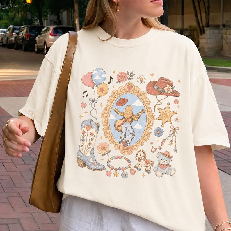 May include: A cream-colored short-sleeve t-shirt featuring a central graphic print of a person playing a guitar inside an ornate gold frame, surrounded by illustrations of a cowboy boot, a hat, a sheriff star, balloons, a teddy bear, and a horse.