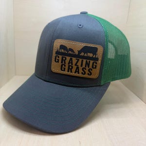 Könnte beinhalten: Grau-grüne Trucker-Cap mit gebogenem Schirm. Die Kappe hat einen braunen Lederaufnäher mit den Worten "GRAZING GRASS" und einer Silhouette von grasenden Tieren. Die Rückseite der Kappe ist aus grünem Mesh.