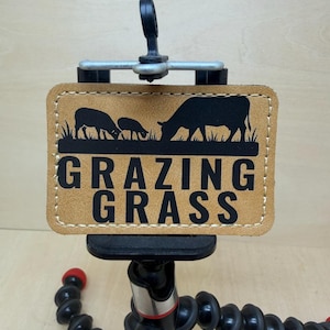 Könnte beinhalten: Ein beigefarbenes Lederpatch mit den Worten "GRAZING GRASS" in fetten schwarzen Buchstaben. Das Patch zeigt eine schwarze Silhouette von Kühen, die auf Gras weiden. Das Patch ist auf einem schwarzen und roten flexiblen Stativ befestigt.