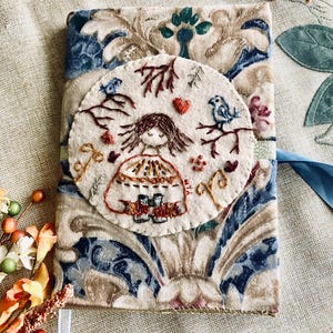 Op de afbeelding: Een rechthoekige omslag voor een dagboek met een blauwe en beige damaststoffen basis en een cirkelvormige, crèmekleurige vilten patch. De patch toont handgeborduurde details van een persoon met een verrekijker, omringd door vogels en takken.