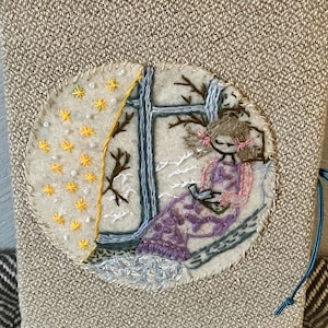 Puede incluir: Un diario de lino beige con un diseño bordado circular que representa a una niña sentada junto a una ventana con un libro. La ventana tiene una cortina amarilla y la escena se desarrolla en un paisaje invernal nevado.