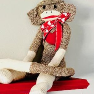 May include: A brown sock monkey wearing a white hat and a red scarf with white hearts. The monkey is sitting on a red shelf.