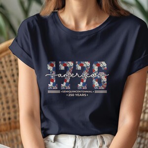 Puede incluir: Camiseta azul marino con el año "1776" en un estampado floral rojo, blanco y azul. La palabra "america" está escrita en cursiva encima del año. Debajo, las palabras "SEMIQUINCENTENNIAL" y "250 YEARS."