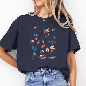 Puede incluir: Camiseta azul marino con un diseño patriótico. El diseño incluye una bandera estadounidense, fuegos artificiales y otras ilustraciones festivas. El texto de la camiseta dice "sparkle with all your might".
