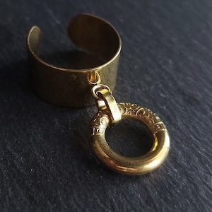 May include: A gold-colored ear cuff with a dangling circular charm. The ear cuff has a simple, open design, while the charm features an ornate, engraved pattern. The jewelry is set against a dark background.