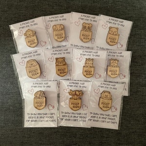 May include: A selection of wooden pocket hug tokens, each with a different animal design, including a monkey, elephant, and panda. Each token is packaged with a card that reads "A Pocket Hug From me to you."