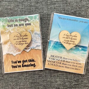 May include: Two cards in plastic sleeves. Each card features a beach scene and a wooden heart with a message. One card reads "Life is tough, but so are you." The other says "Always remember, you are: BRAVER..."