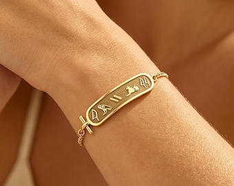 Puede incluir: Una pulsera de tono dorado con un colgante de barra rectangular de extremos redondeados. La superficie de la barra está grabada con una serie de símbolos jeroglíficos egipcios distintos. La pulsera se asegura con una cadena fina y un cierre de barra.