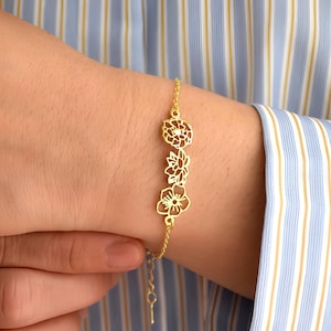 May include: A gold bracelet with a delicate floral design. The bracelet features three cutout flowers in a row, each with intricate details. The bracelet is adjustable and has a lobster clasp closure.
