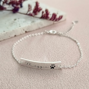 Puede incluir: Un brazalete de cadena de plata con un dije de barra personalizado. El dije está grabado con el nombre "Maggie" y una huella de pata.