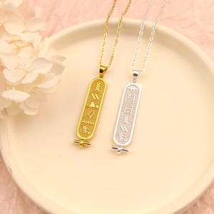 Könnte beinhalten: Zwei Anhängerhalsketten mit ägyptischem Hieroglyphen-Design. Eine Halskette ist goldfarben, die andere silberfarben. Beide Anhänger sind rechteckig mit eingravierten Symbolen und hängen an filigranen Ketten. Die Halsketten liegen auf einem weißen Teller.