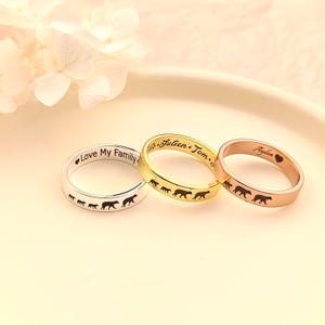 May include: Three personalised rings with a design of a family of bears walking. The rings are silver, gold, and rose gold. The silver ring says "Love My Family", the gold ring says "Julien Tom", and the rose gold ring says "Ayden".
