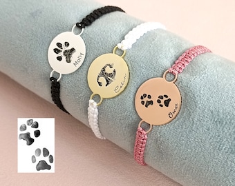 May include: Three personalized dog paw print bracelets with black, white, and pink cords. The silver bracelet says "Moin", the gold bracelet says "Celia", and the rose gold bracelet says "Oscar".