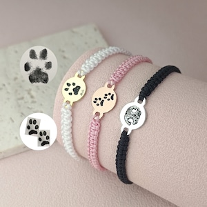 May include: Three bracelets with paw print charms. The bracelets are white, pink, and black. The charms are silver, gold, and rose gold.