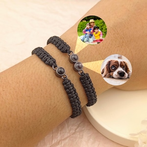 Könnte beinhalten: Zwei schwarze geflochtene Armbänder mit dunkelgrauen Akzenten werden an einem Handgelenk gezeigt. Jedes Armband hat einen kreisförmigen Anhänger und dunkelgraue Perlen. Der Hintergrund enthält zwei kreisförmige Bilder: eine Familie und ein Hund.