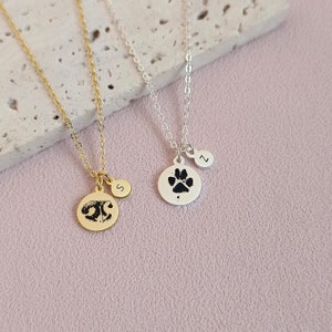Könnte beinhalten: Zwei goldene und silberne Halsketten mit Pfotenabdruck-Anhängern. Die goldene Halskette hat einen kleinen Anhänger mit dem Buchstaben "S" und die silberne Halskette hat einen kleinen Anhänger mit dem Buchstaben "Z".