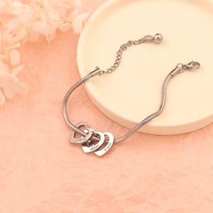 May include: Silver snake chain bracelet with three interconnected heart charms. Each heart charm is engraved with a name: Cheryl, Allison, and Paul.