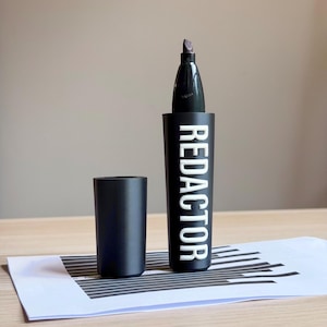 May include: A black marker with a removable cap, featuring the word REDACTOR printed in bold white capital letters on the barrel. The marker has a thick, dark tip and a textured matte finish on the body, standing upright next to its detached cap.