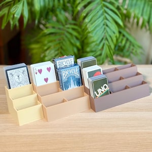 May include: Three-tiered card holders in beige, tan, and brown, each with three compartments. The holders contain playing cards and decks of cards, including Bicycle and Uno brands. A green leafy plant is in the background.