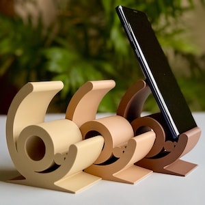 May include: Three beige tape dispensers with a black smartphone holder. The dispensers are shaped like a snail shell and have a beige roll of tape inside.