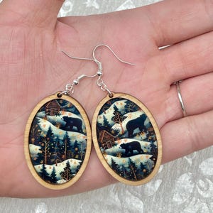 Pu&ograve; includere: Orecchini ovali in legno con una scena invernale che presenta una capanna, alberi e orsi. Gli orecchini sono dipinti in tonalit&agrave; di blu, verde e bianco con accenti dorati.