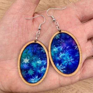 Pode incluir: Brincos ovais de madeira com um design de floco de neve azul e branco. Os brincos t&ecirc;m ganchos prateados.