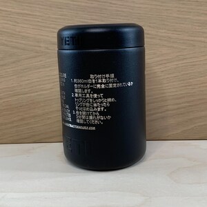 May include: A black YETI insulated container with a screw-on lid. The container has text in Japanese on the side. The container is cylindrical and sits on a light-colored wooden surface.