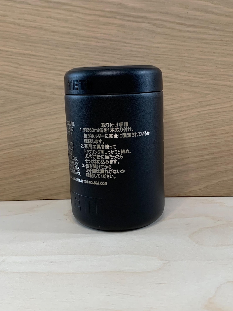 May include: A black YETI insulated container with a screw-on lid. The container has text in Japanese on the side. The container is cylindrical and sits on a light-colored wooden surface.