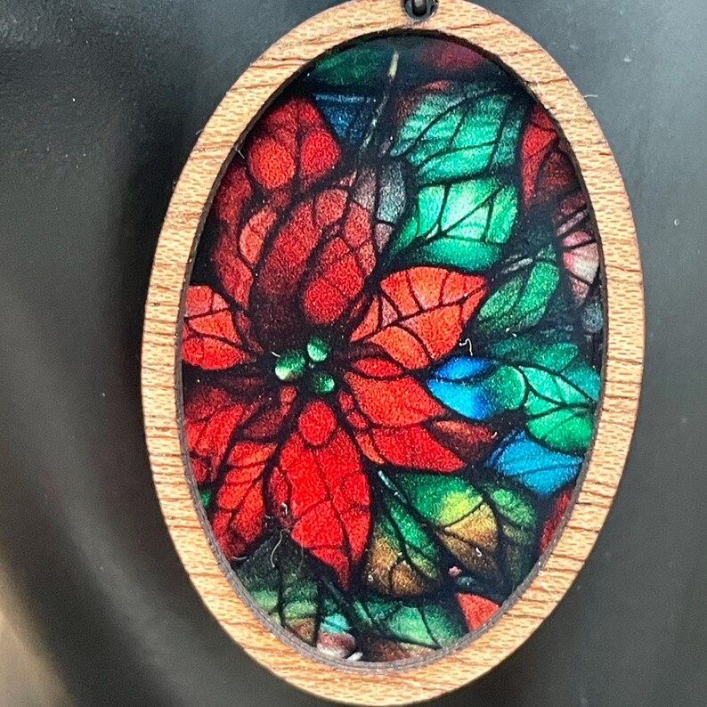 May include: A wooden oval ornament featuring a stained glass style image of a red poinsettia flower with green leaves.