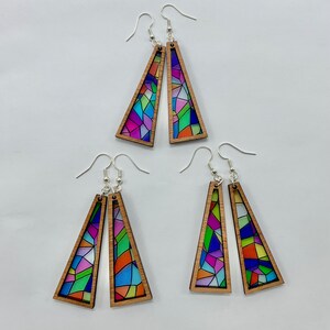 May include: A set of three pairs of colorful stained glass earrings. Each pair features a geometric design with a variety of colors, including blue, green, purple, and orange. The earrings are made of wood and have silver hooks.