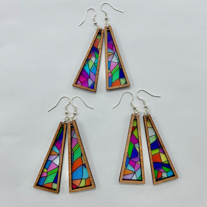 May include: A set of three pairs of colorful stained glass earrings. Each pair features a geometric design with a variety of colors, including blue, green, purple, and orange. The earrings are made of wood and have silver hooks.