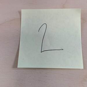 May include: A light green sticky note with the number "2" written in black ink. The note is rectangular and slightly angled, with the number centered on the paper. The background is a light brown wooden surface.