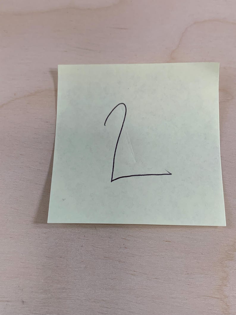 May include: A light green sticky note with the number "2" written in black ink. The note is rectangular and slightly angled, with the number centered on the paper. The background is a light brown wooden surface.
