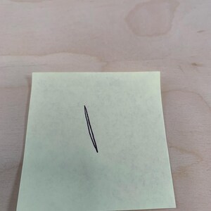 May include: A yellow sticky note with a black, diagonal line drawn on it. The note is rectangular and slightly angled, with the line extending from the top to the bottom. The background is a light-colored wooden surface.