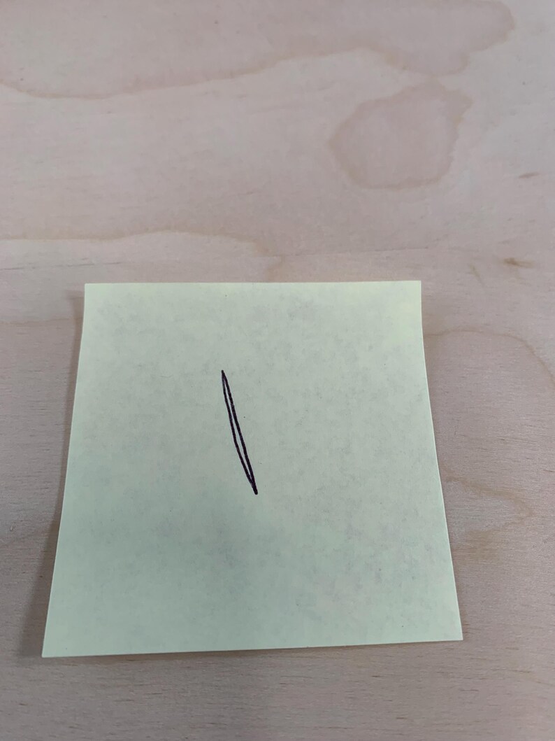 May include: A yellow sticky note with a black, diagonal line drawn on it. The note is rectangular and slightly angled, with the line extending from the top to the bottom. The background is a light-colored wooden surface.