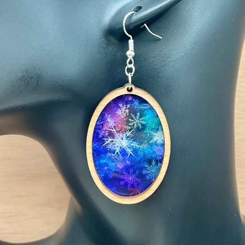 Pode incluir: Brincos ovais de madeira com estampa de gal&aacute;xia azul, roxo e rosa com flocos de neve brancos.