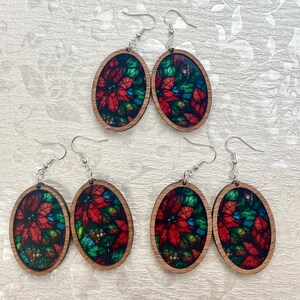 May include: Four pairs of oval-shaped wooden earrings with a stained glass design featuring red poinsettia flowers and green leaves. The earrings have silver hooks.