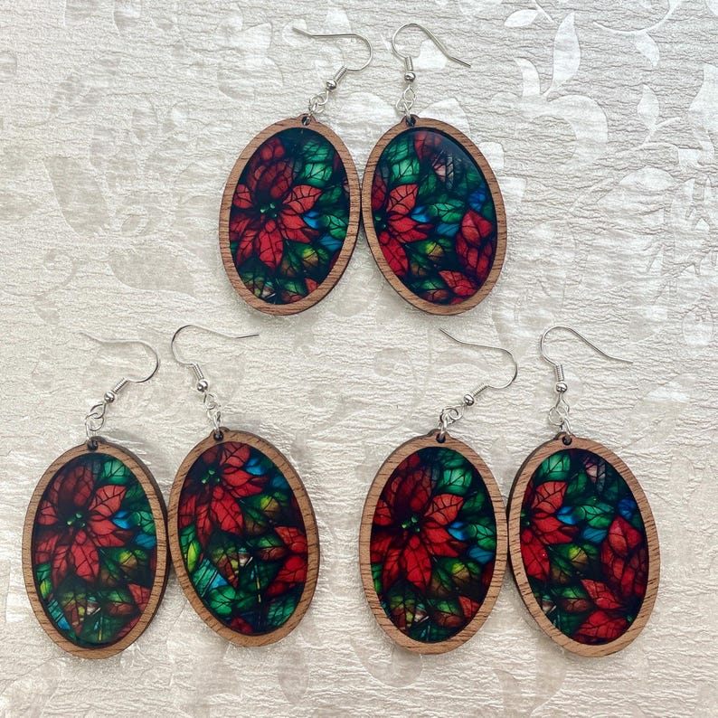 May include: Four pairs of oval-shaped wooden earrings with a stained glass design featuring red poinsettia flowers and green leaves. The earrings have silver hooks.