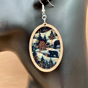 Pu&ograve; includere: Orecchini ovali in legno con una scena invernale che presenta una capanna, alberi e due orsi neri che camminano nella neve. Lo sfondo &egrave; blu scuro con accenti dorati.