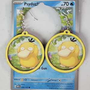 May include: A pair of wooden earrings featuring a yellow duck character from the Pokemon franchise. The earrings are circular and have a yellow border. The duck is depicted with a surprised expression and is holding its head with its hands. The earrings are hanging from silver hooks.