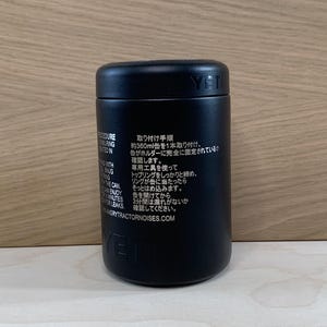 May include: Black cylindrical container with the brand name "YETI" printed on the side. The container has text in English and Japanese. The container is on a light wood surface.
