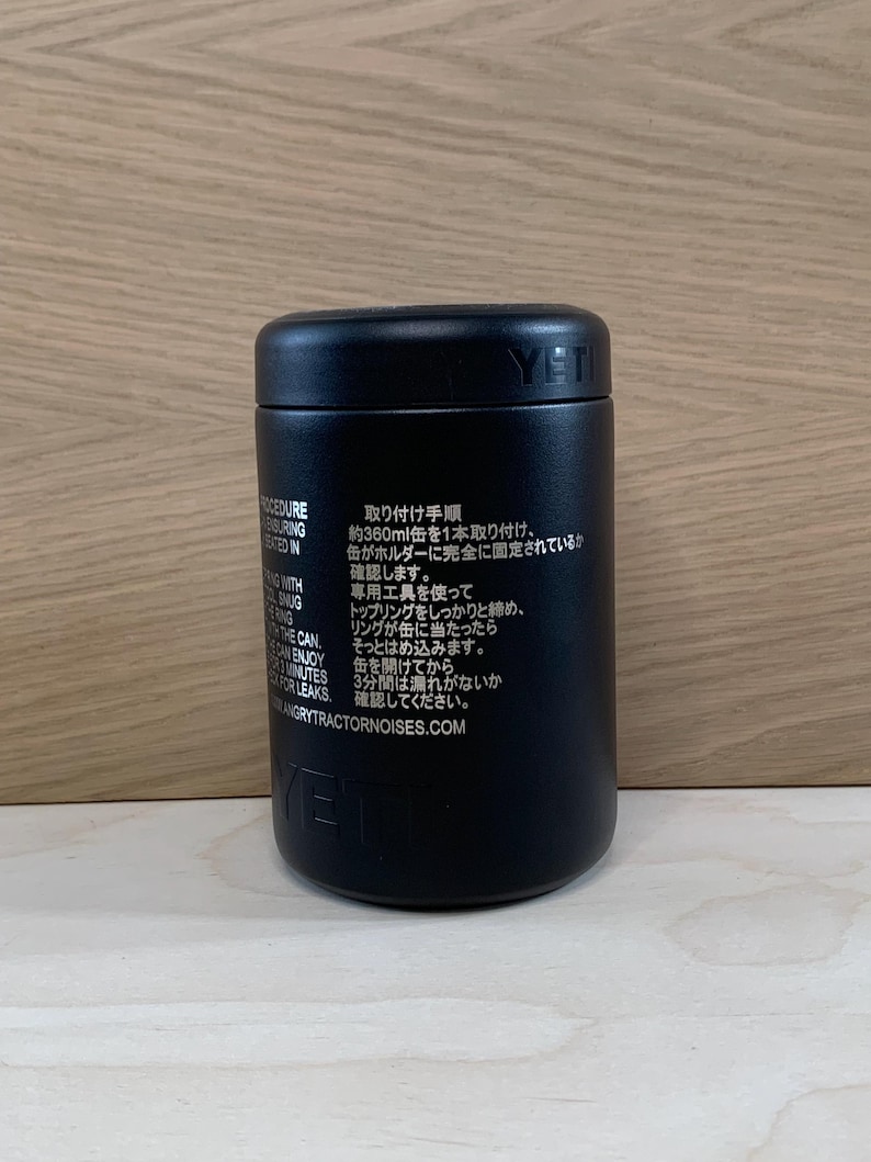 May include: Black cylindrical container with the brand name "YETI" printed on the side. The container has text in English and Japanese. The container is on a light wood surface.